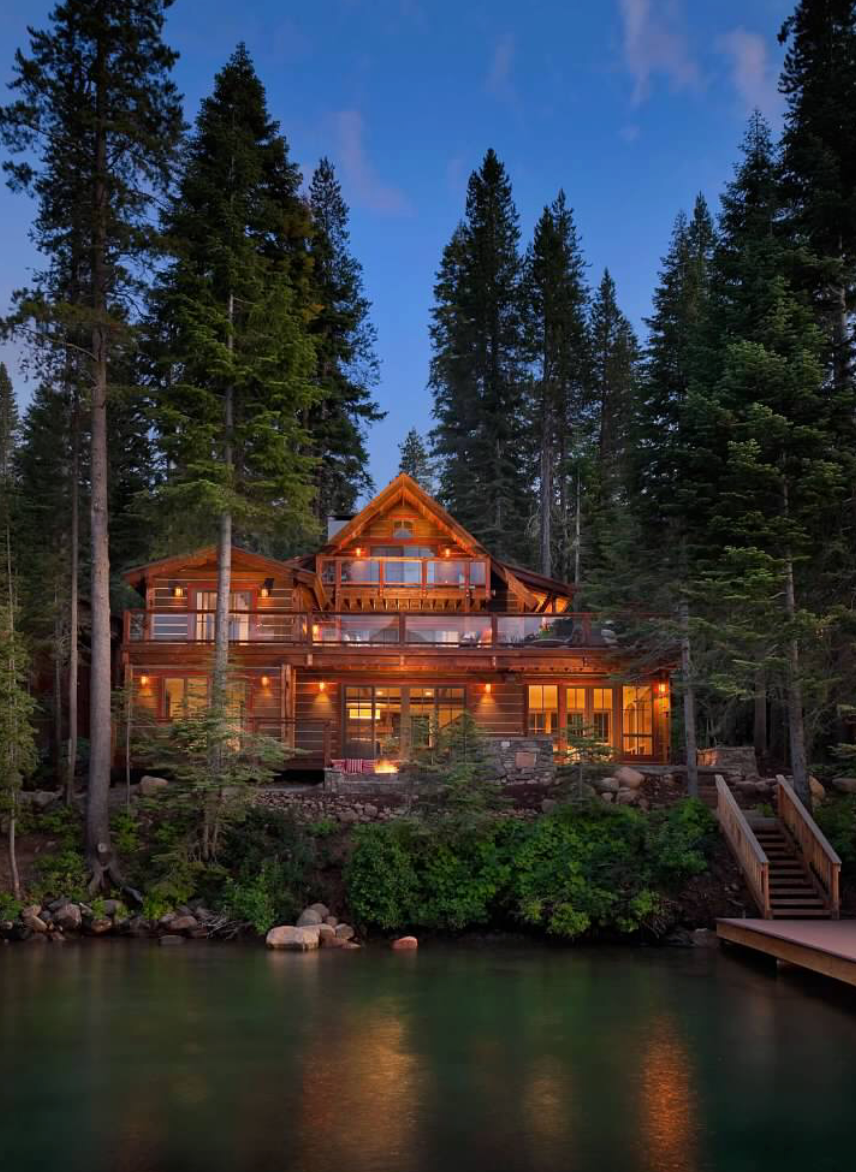 Luxury cabin surrounded by tall trees by a lake at night.