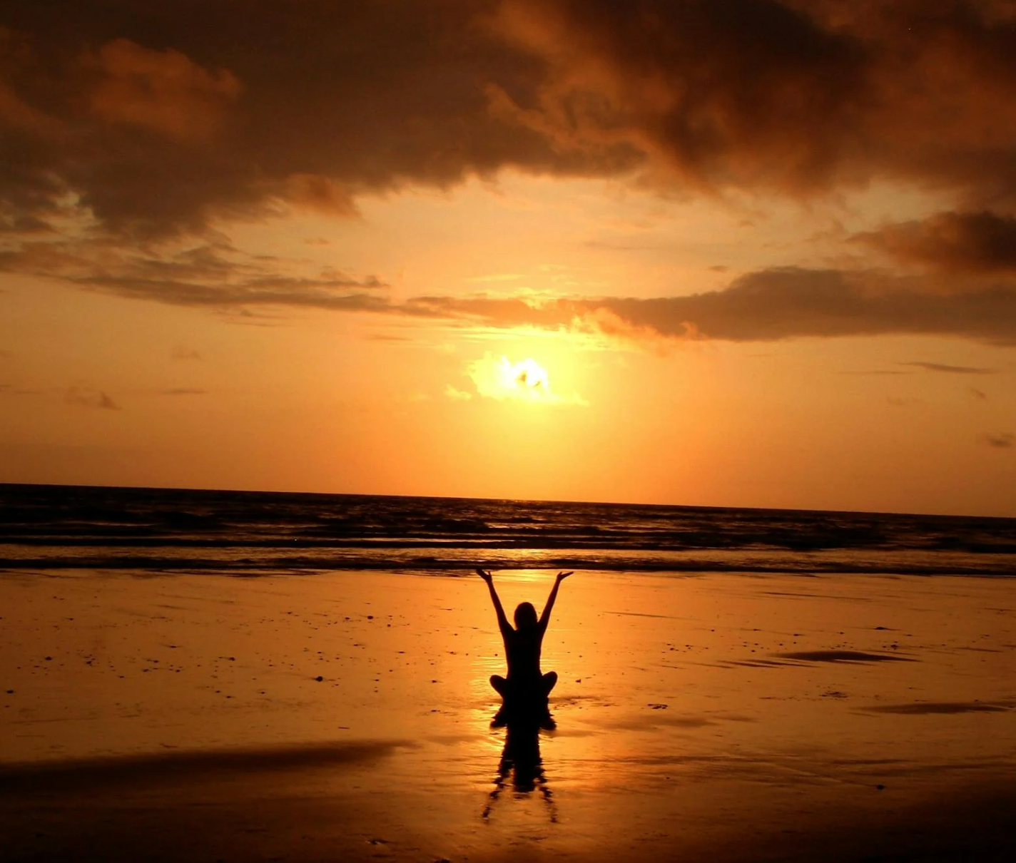 beach with sun setting shows woman sitting on the sand saluting the sun