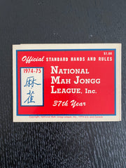 A vintage 1914-1975 official standard hands and rules card for Mah Jongg, featuring a red background with blue and white borders and text, photographed on a dark textured surface.