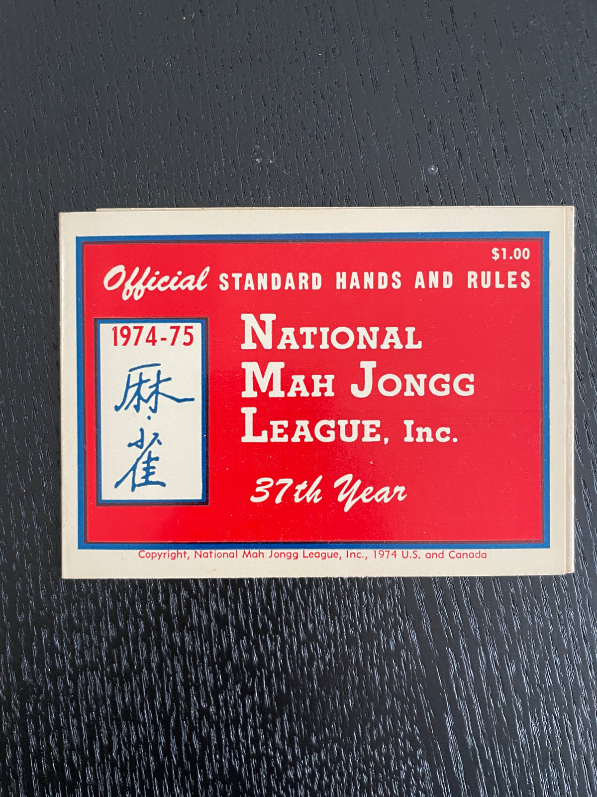 A vintage 1914-1975 official standard hands and rules card for Mah Jongg, featuring a red background with blue and white borders and text, photographed on a dark textured surface.