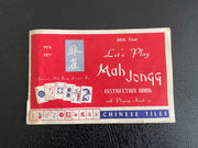 A vintage Chinese mahjong instruction book cover, featuring a red background with white and blue text and illustrations of playing cards, photographed flat against a dark textured surface.